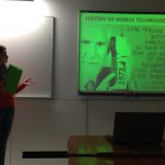 Studentka stojąca w sali wykładowej przy tablicy na której wyświetlana jest prezentacja multimedialna