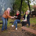 Trzy studentki na ławkach w otoczeniu drzew jedzą kiełbasę z ogniska