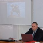 Dr Konstantin Stankov z uczelni w Bułgarii siedzi przy biurku, przed nim otwarty laptop w tle na ekranie prezentacja multimedialna