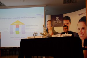 Jeden z prelegentów przy stole prezydialnym, obok ekran z prezentacją multimedialną