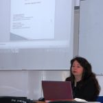 Dr Rumyana Angelowa siedzi przy biurku, przed nią otwarty laptop, za plecami ekran z wyświetloną prezentacją towarzyszącą wykładowi