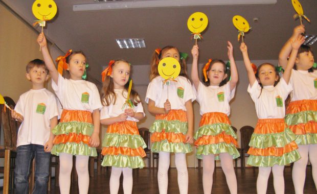 Dzieci (sześć dziewczynek i jeden chłopiec) w trakcie przedstawienia. Dziewczynki mają białe podkoszulki i zielono pomarańczowe spódniczki, unoszą w górę żółty znaczek z uśmiechniętą buzią