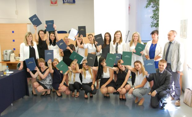 Zdjęcie grupowe studentów po egzaminie dyplomowym, studenci prezentują przed sobą prace dyplomowe, pierwszy rząd kuca