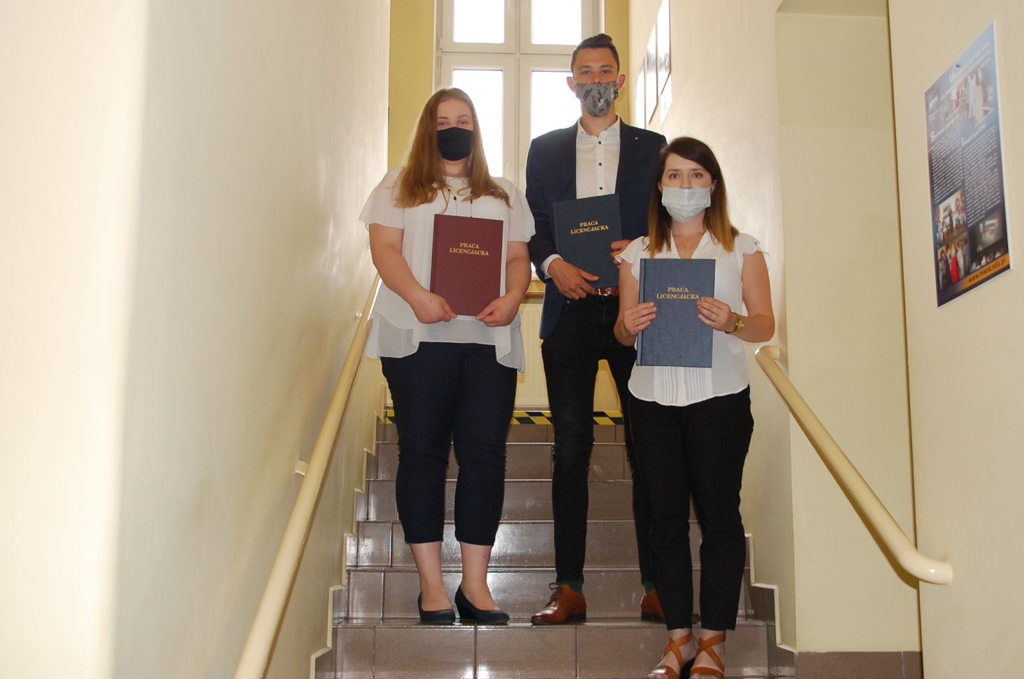 Troje studentów pozuje z pracami dyplomowymi na schodach budynku przy Szerokiej 9